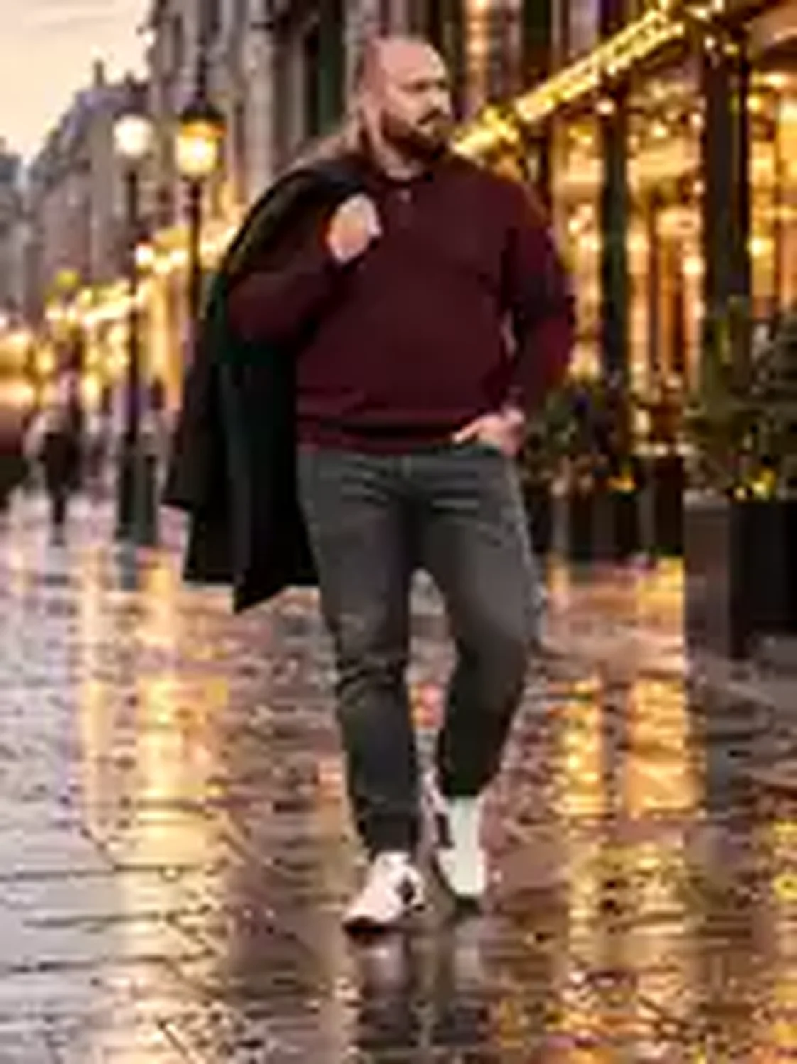 Kräftiger Mann in bordeauxfarbenem Half-Zip-Pullover, dunkler Jeans, rotweißen Sneakers und schwarzem Mantel über der Schulter bei Abendlicht.
