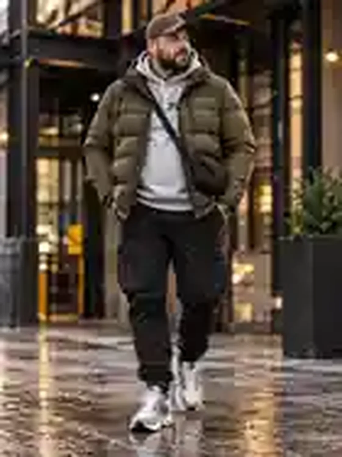 Stämmiger XXL-Mann mit olivgrüner Pufferjacke, grauem Hoodie, schwarzer Cargohose, grau-weißen Sneakers und Crossbody-Bag im Cityviertel.